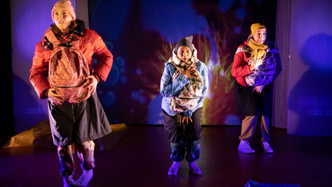Tre Skådespelare på en teaterscen med tjocka vinterkläder och varsin ryggsäck på magen.