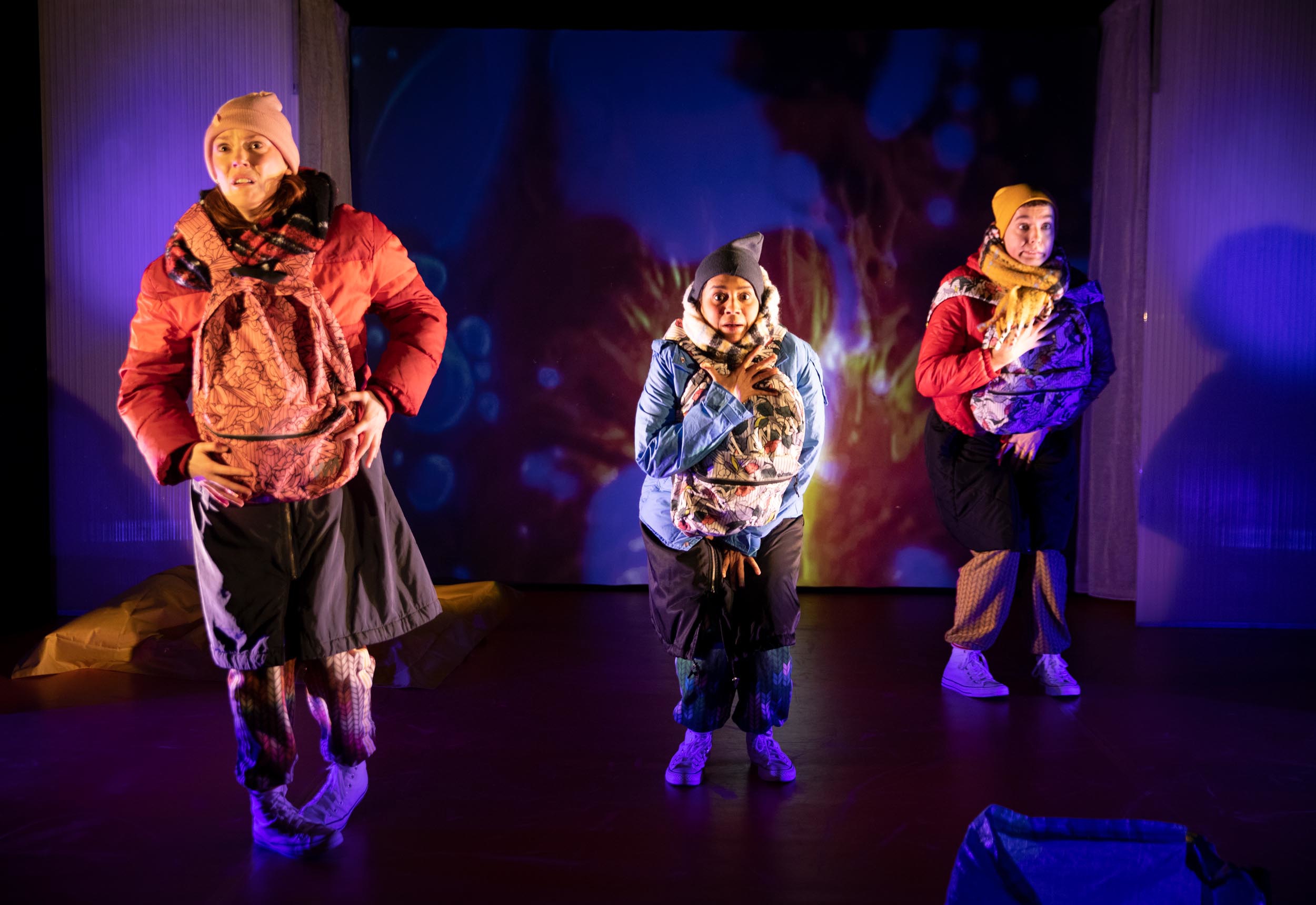 tre skådespelare på en teaterscen med tjocka vinterkläder och varsin ryggsäck på magen.