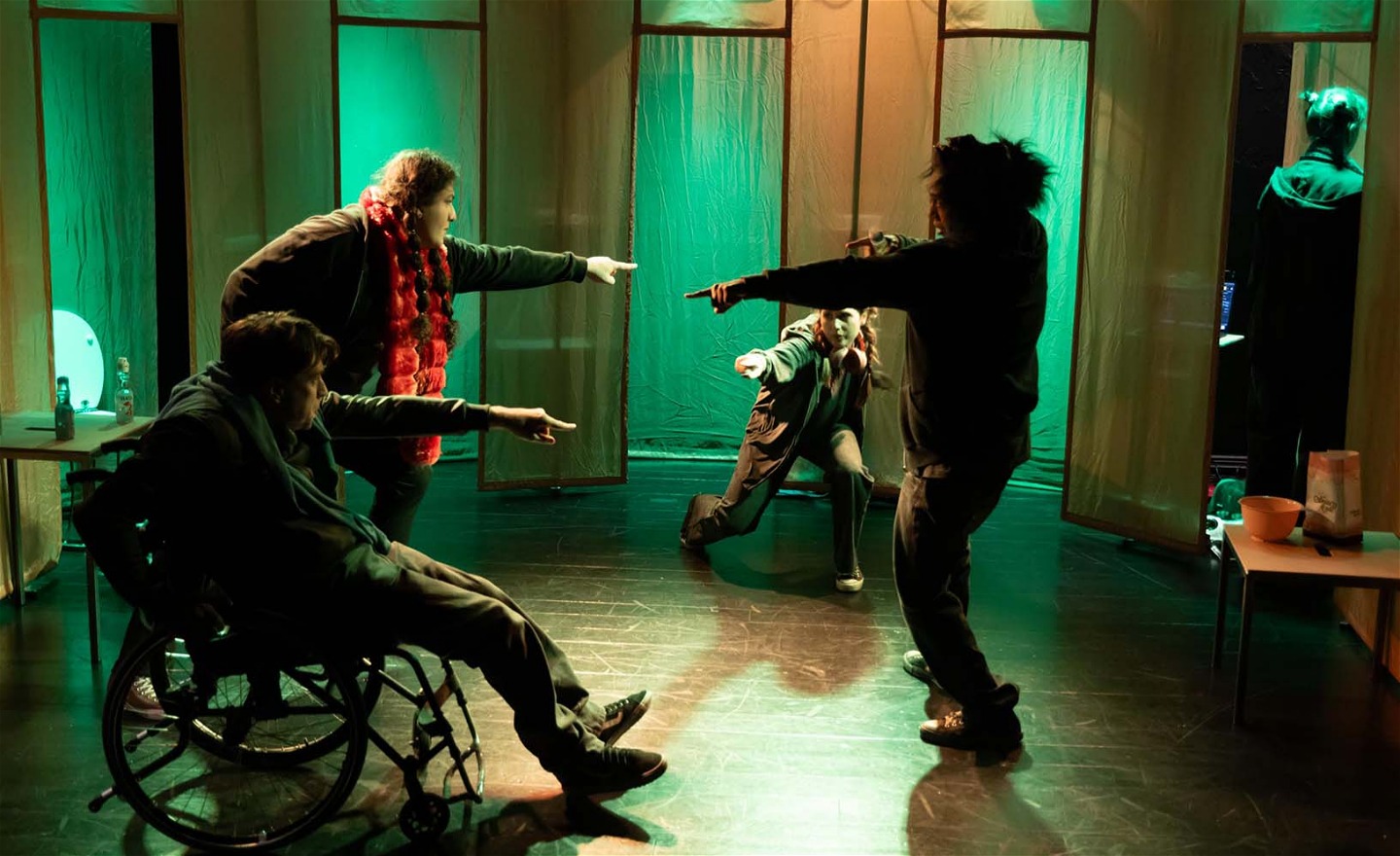 Bild från föreställningen. Fyra personer står i en ring och pekar på varandra. En sitter i rullstol och en står med böjda ben.